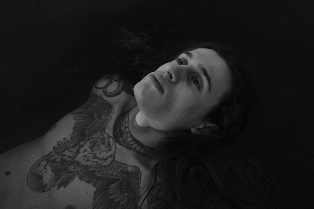 Monochrome portrait of a tattooed man with long hair, lying down, evoking solitude.