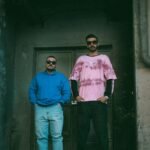 Two men in casual urban wear standing confidently against a gritty backdrop.