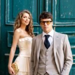 A stylish couple dressed in formal attire stands in front of a rustic, teal-painted door.